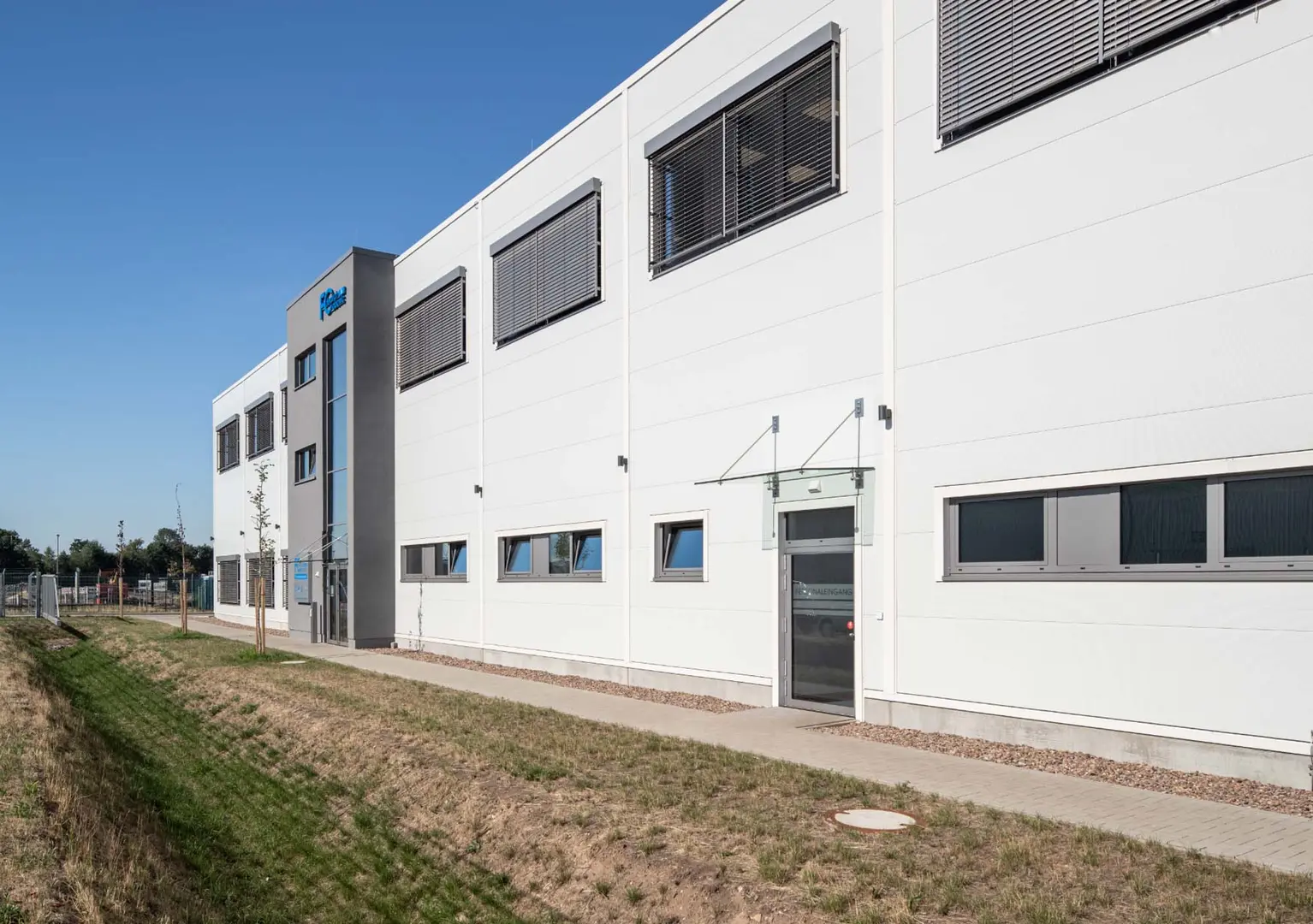Dargestellt ist ein großes Gebäude, das als Lager- und Fertigungshalle sowie Bürotrakt dienen soll. Hierbei sind ein Personaleingang und ein Haupteingang zu sehen, sowie diverse Fenster im ersten Stock und im Erdgeschoss.
