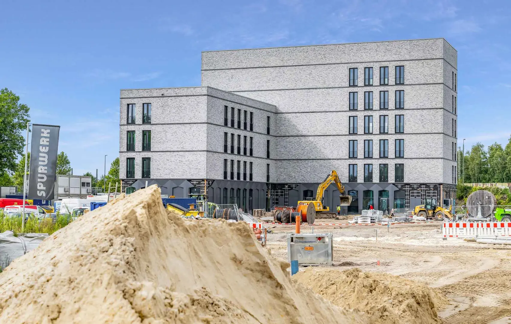 Fotografie des Bauprozesses des Spurwerkkomplexes. Dargestellt ist ein fertiges großes Gebäude aus grauem Stein, davor ein Bagger auf einem großen aufgeschütteten Feld.