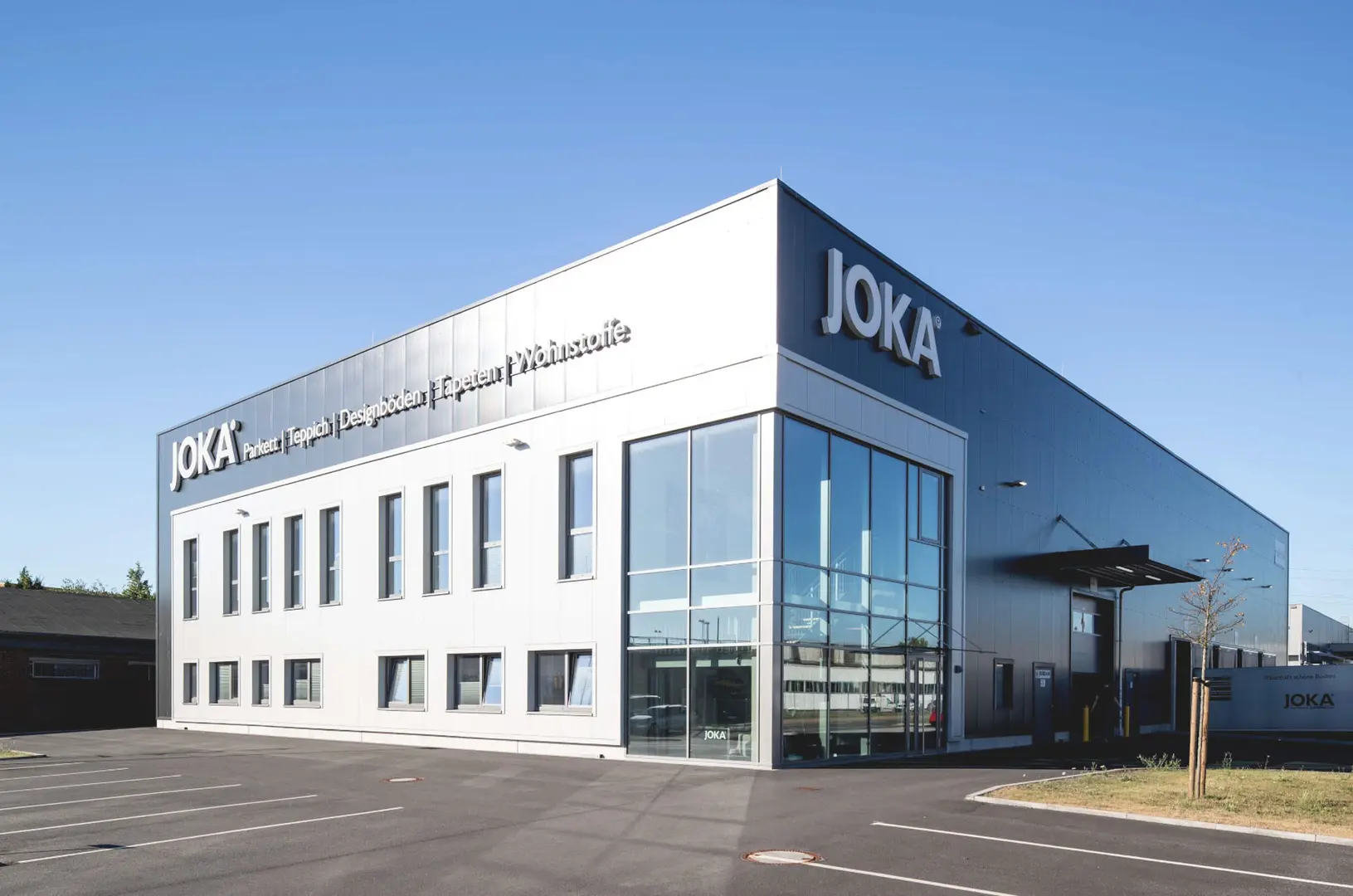 Foto einer großen Halle mit der Aufschrift "JOKA". Dargestellt ist eine große, dunkle Halle mit einer verglasten Eckfront und einer Ladeluke für LKW. Oben unter dem Dach steht "JOKA, Paket, Teppich, Designböden, Tapeten, Wohnstoffe".