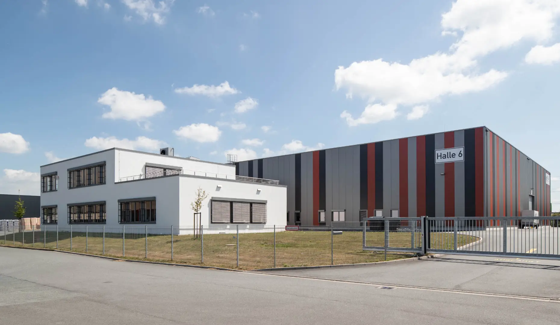 Foto der Halle 6 mit angrenzendem Bürogebäude. Abgebildet ist eine große Halle mit der Aufschrift "Halle. 6", davor ein kleineres weißes Gebäude, das als Bürogebäude dient.